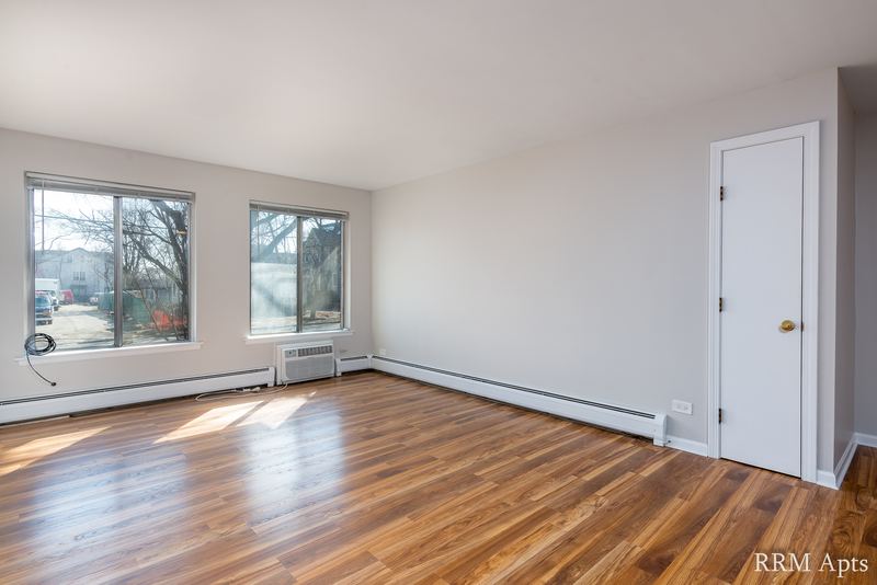 Living room with wood floor, two windows, a wall air conditioner and a white entry door, 4th and Broadway apartment 1B