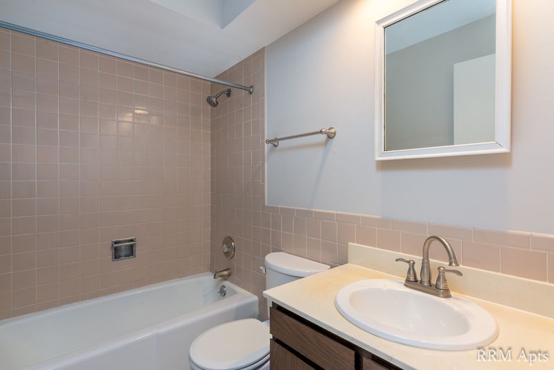Bathroom with beige tile tub surround, toilet, dark wood vanity with white sink and mirror, 4th and Broadway apartment 1B