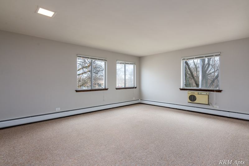 Carpeted living room with two windows, baseboard radiator and a wall air conditioner, 4th and Broadway apartment 2A