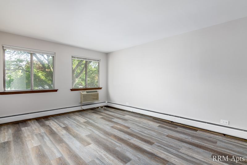 Living room with gray wood-look floor, two windows, wall air conditioner and radiator, 4th and Broadway apartment 2B