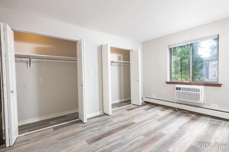 Bedroom with gray wood-look floor, two open bifold closets and a wall air conditioner, 4th and Broadway apartment 2B