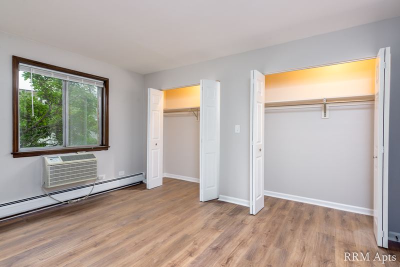 Bedroom with wood floor, window with wall air conditioner and two open bifold closets, 4th and Broadway apartment 2C