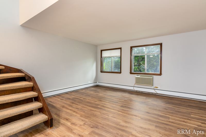 Living room with wood floor, two windows and carpeted stairs leading up on the left, 4th and Broadway apartment 2C