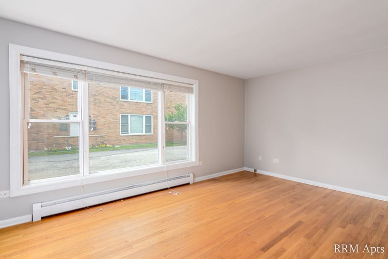 Living room with wood floor, a wide window facing a brick building and a baseboard radiator, Austin Avenue apartment 118 A