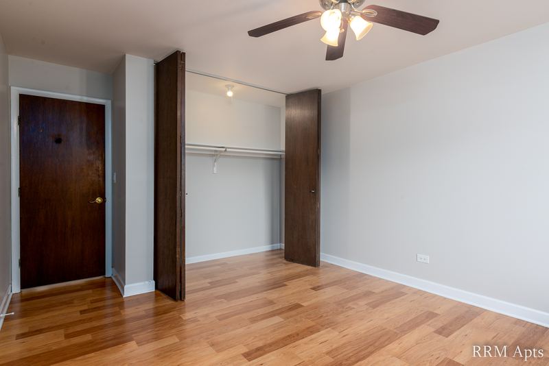 Bedroom with wood floor, ceiling fan, wood door on the left and an open closet with dark doors, Cook Street apartment 4