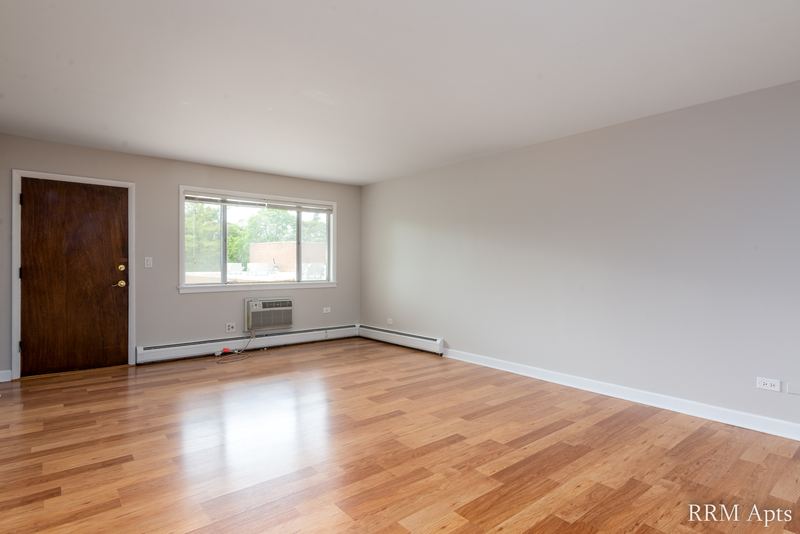 Living room with wood floor, wide window with a wall air conditioner below and a wood entry door, Cook Street apartment 4