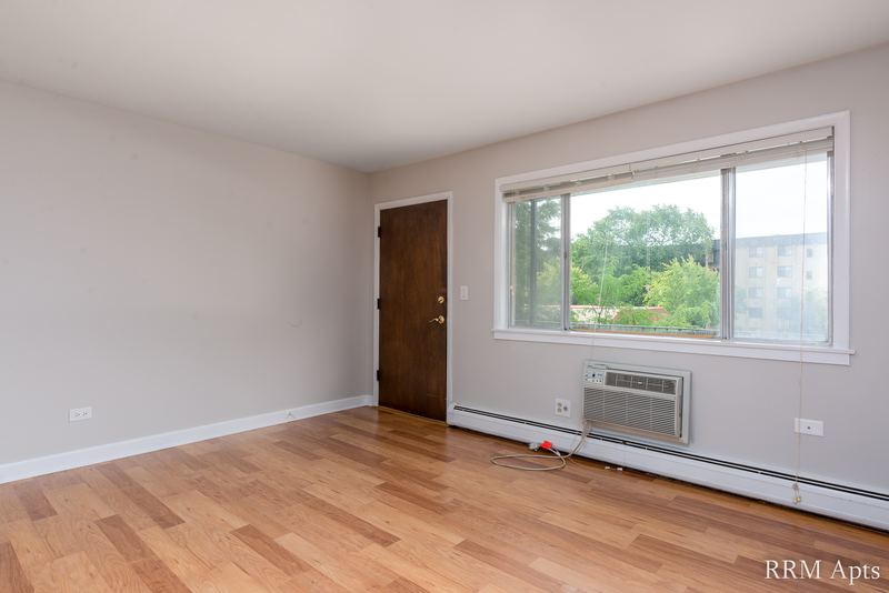 Living room with wood floor, a wide window facing a brick building and a wall air conditioner, Cook Street apartment 4