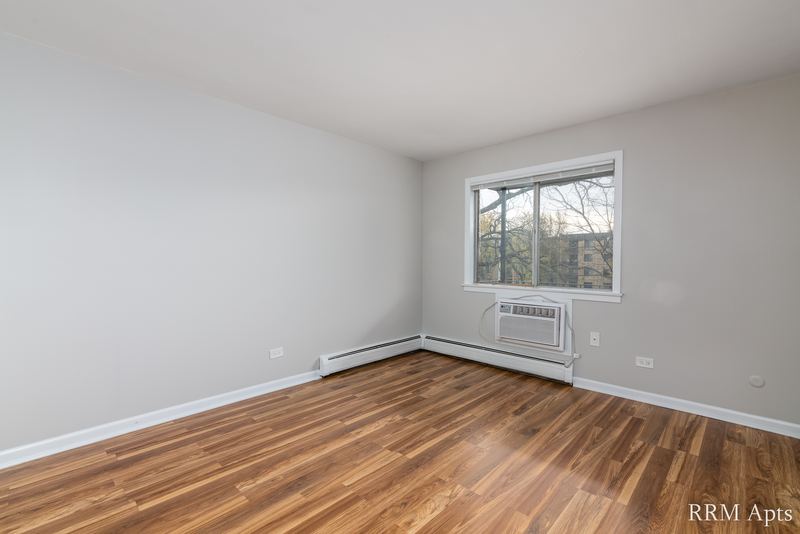 Living room with wood floor, wide window with a wall air conditioner below and baseboard radiator, Cook Street apartment 9