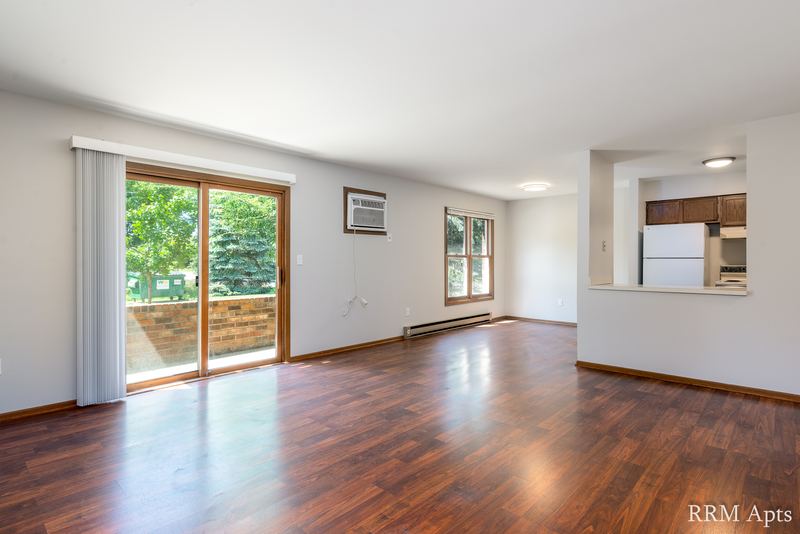 Living room with wood floor, sliding glass door to patio, wall air conditioner and kitchen beyond, Depot Road apartment 1