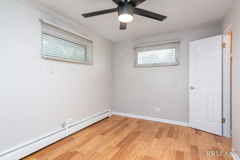 Bedroom with wood floor, ceiling fan, two windows with blinds and a white door, East Park apartment 3