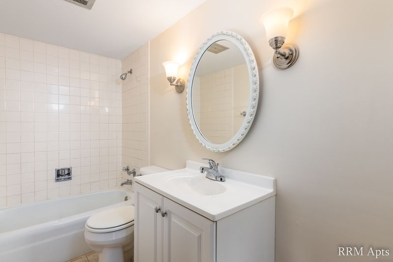 Bathroom with white tile tub surround, toilet, white vanity and an oval mirror with sconces, John Mogg apartment 32927 203