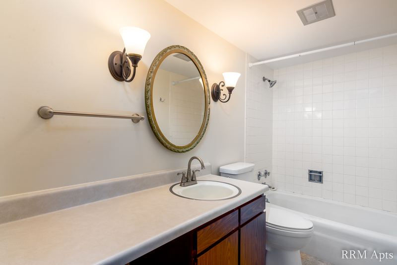 Bathroom with white tile tub surround, toilet, long dark wood vanity and an oval mirror, John Mogg apartment 32927 301
