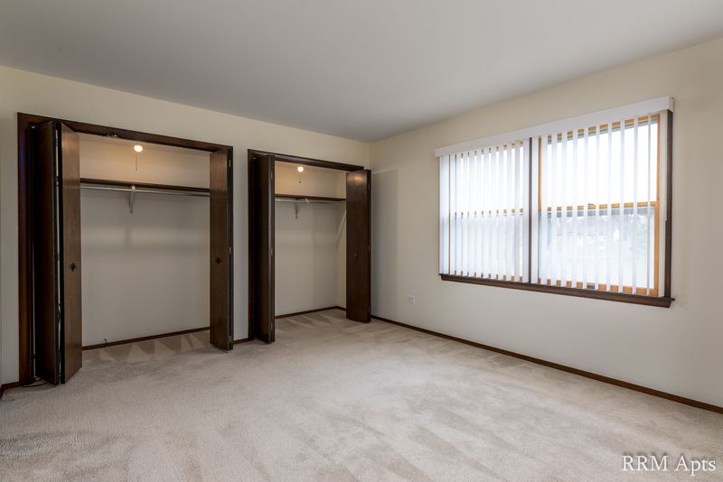 Carpeted bedroom with two open closets with dark wood doors and a window with blinds, John Mogg apartment 32927 304