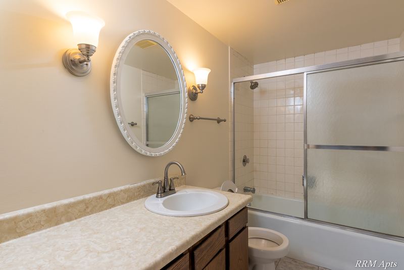 Bathroom with sliding glass tub doors, oak vanity with beige counter and an oval mirror, John Mogg apartment 32985 201