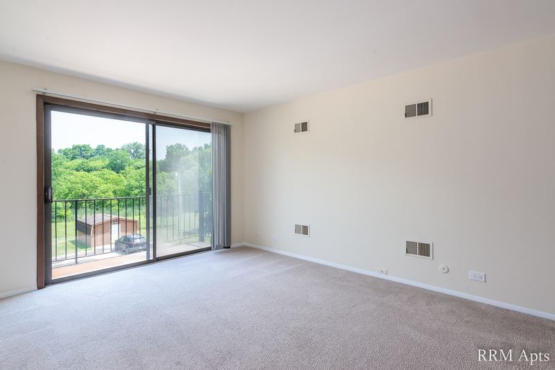 Carpeted living room with sliding glass door to a balcony with iron railing and trees beyond, John Mogg apartment 32985 301
