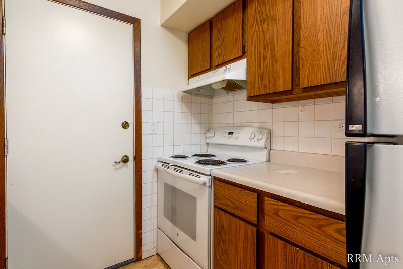 Kitchen with white electric stove, oak cabinets, black refrigerator and a white door, John Mogg apartment 32985 301