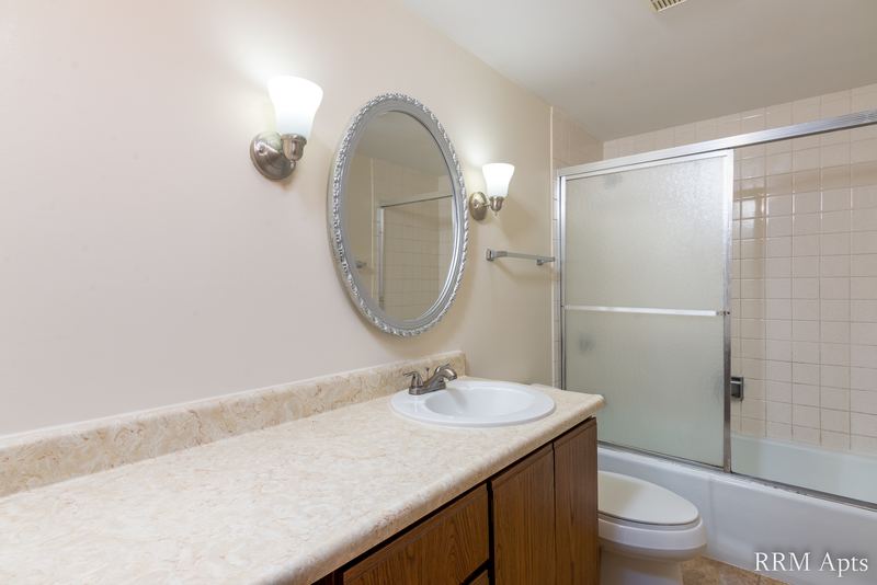 Bathroom with sliding glass tub doors, toilet, oak vanity and an oval mirror with sconces, John Mogg apartment 32985 301