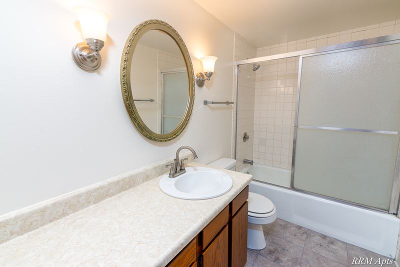 Bathroom with wood vanity, oval mirror, sconce lights, sliding glass tub door and white tile, John Mogg apartment 32985 304 N