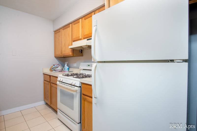 Kitchen with wood cabinets, white stove under a range hood, refrigerator and beige tile floor, Maple Avenue apartment 1B