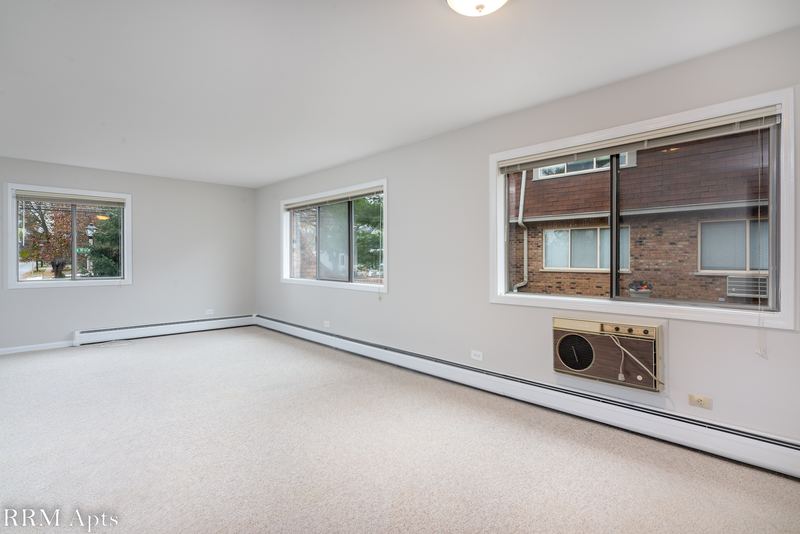 Living room with beige carpet, three windows facing a brick building and a wall air conditioner, Maple Avenue apartment 1E