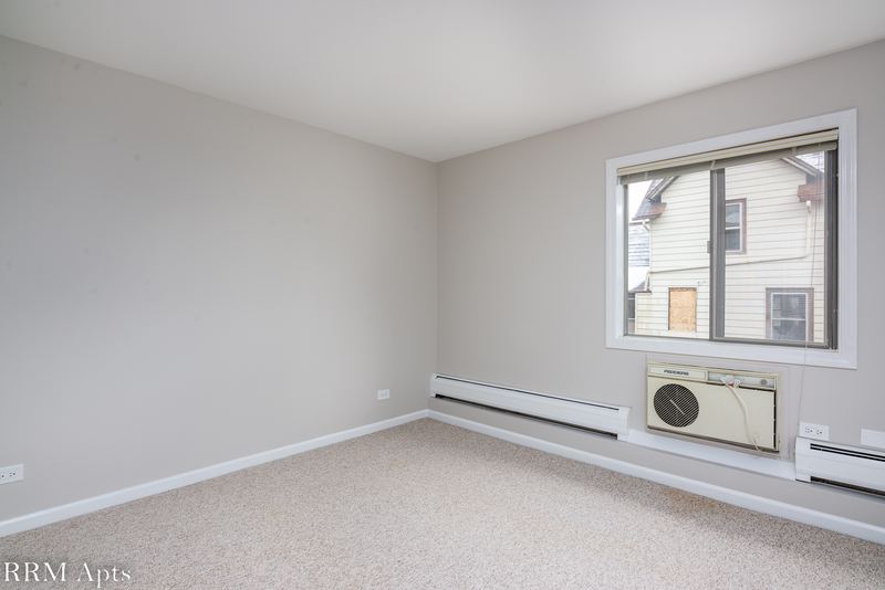Bedroom with beige carpet, a window with wall air conditioner looking out on houses, Maple Avenue apartment 1E