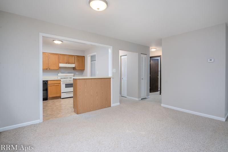Living room with beige carpet, open kitchen with wood cabinets and a hallway to the bedrooms, Maple Avenue apartment 1E