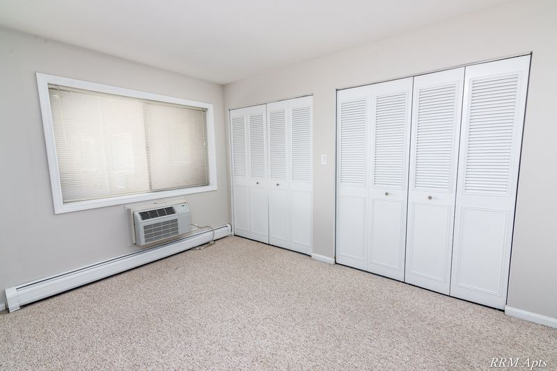Bedroom with beige carpet, a window with wall air conditioner and two white louvered closets, Maple Avenue apartment 1J