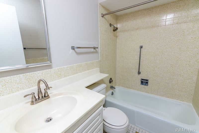 Bathroom with beige tile tub surround, white vanity, mirror and grab bars, Maple Avenue apartment 1J