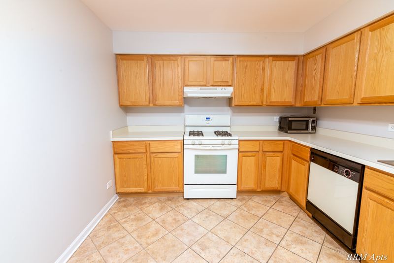 Kitchen with wood cabinets on two walls, white stove and dishwasher, and beige tile floor, Maple Avenue apartment 1J