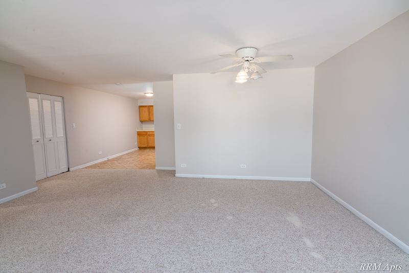 Living room with beige carpet, ceiling fan and a view through to the kitchen with wood cabinets, Maple Avenue apartment 1J