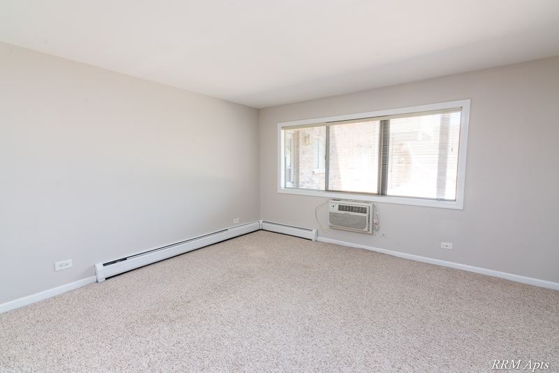 Living room with beige carpet, a wide window with wall air conditioner and baseboard heat, Maple Avenue apartment 1J