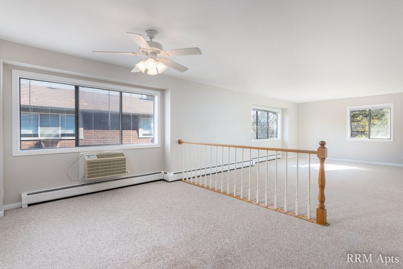 Living room with beige carpet, ceiling fan, wall air conditioner and a white stair railing, Maple Avenue apartment 2A
