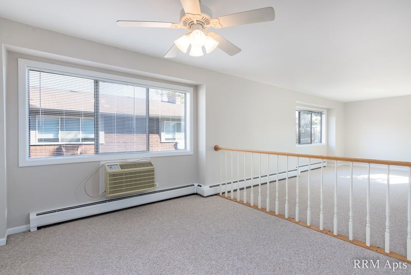 Living room with beige carpet, ceiling fan, two windows and white stair railing, Maple Avenue apartment 2A