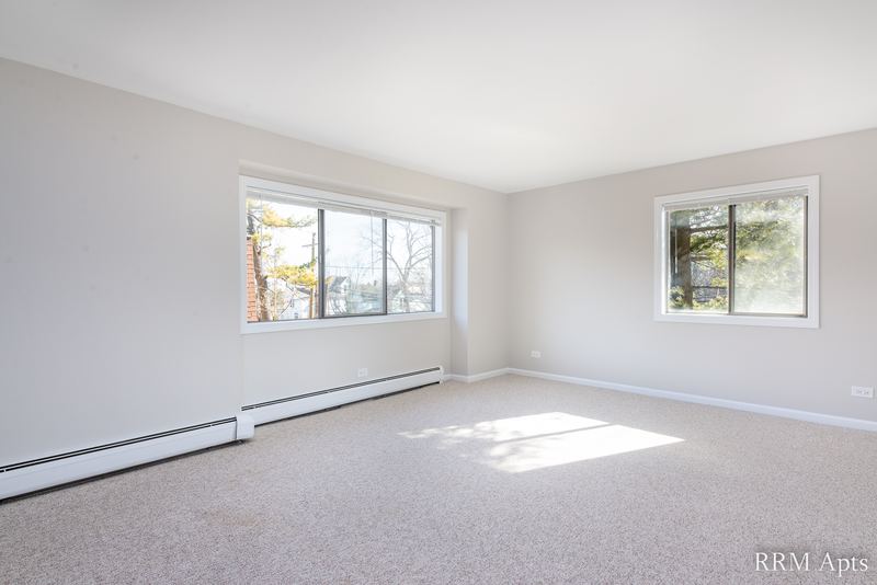 Living room with beige carpet, gray walls, two windows with trees outside and baseboard heat, Maple Avenue apartment 2A