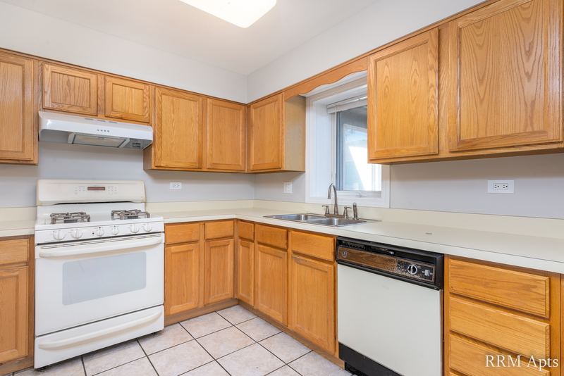 Kitchen with wood cabinets, white gas stove and dishwasher, window over the sink and tile floor, Maple Avenue apartment 2A