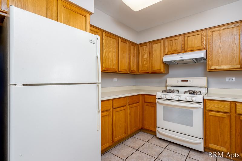 Kitchen with white refrigerator, wood cabinets, white gas stove under a range hood and tile floor, Maple Avenue apartment 2A