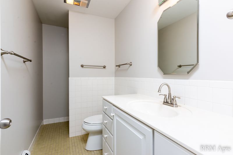 Bathroom with white vanity, mirror, beige tile wainscot, towel bars and toilet, Maple Avenue apartment 2A
