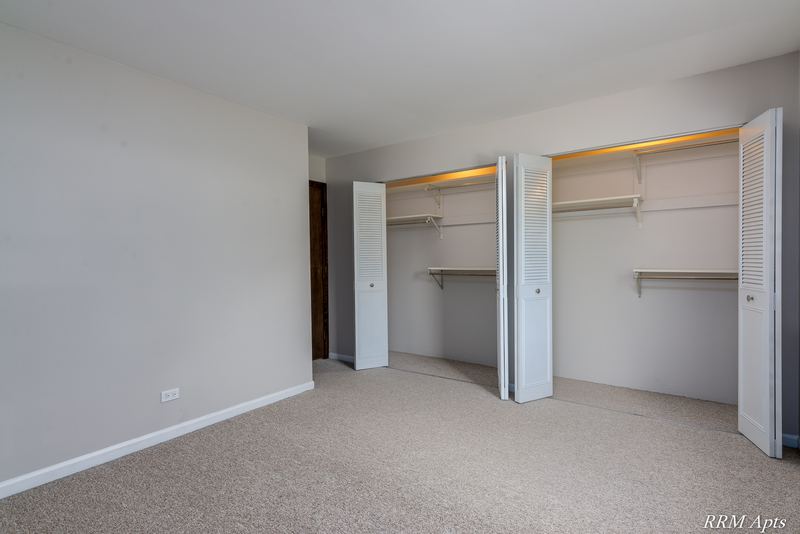 Bedroom with beige carpet, two open closets with white bifold doors and a white door, Maple Avenue apartment 2C