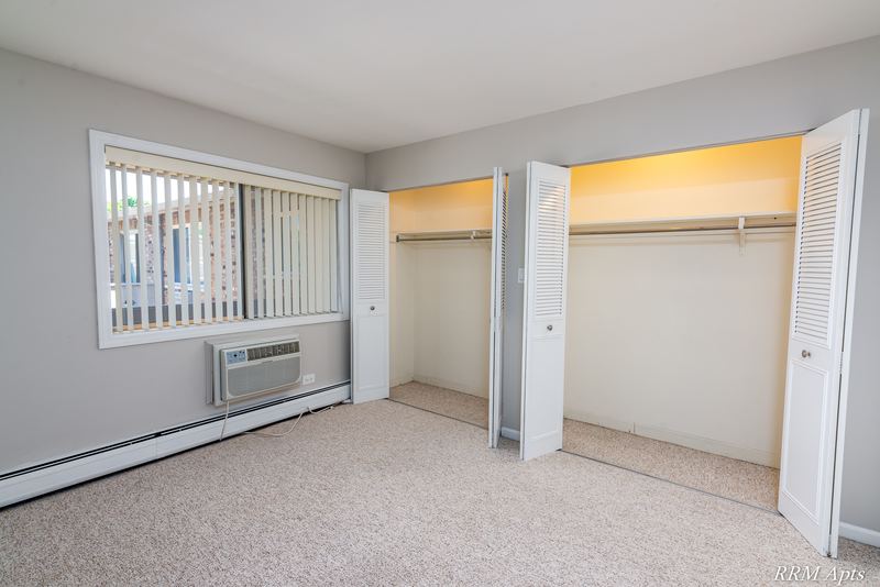 Bedroom with beige carpet, vertical blinds, wall air conditioner and two open closets, Maple Avenue apartment 2J