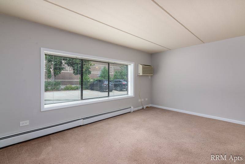 Living room with beige carpet, wide window looking out on parked cars and wall air conditioner, Maple Avenue apartment LL2