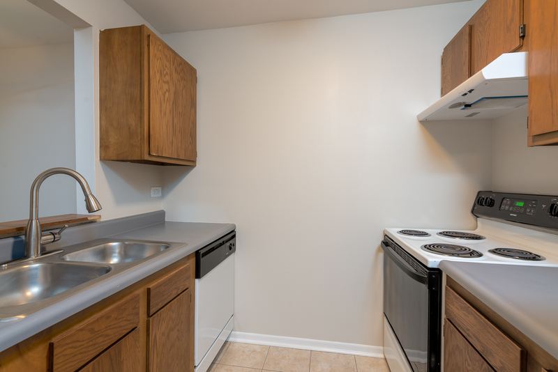 Kitchen with wood cabinets, stainless sink, black dishwasher and electric stove under a range hood, Oriole Court apartment 1C