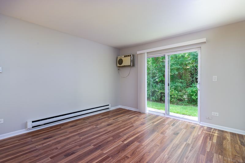 Living room with wood laminate floor, sliding glass door with trees outside and baseboard heat, Oriole Court apartment 1C