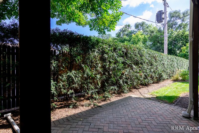 Patio with brick pavers, tall hedges, trees and a strip of lawn seen from the doorway, Park West apartment 613 B