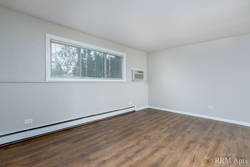 Living room with wood-look floor, wide window with trees and a wall air conditioner, Pioneer Court apartment 900 1D