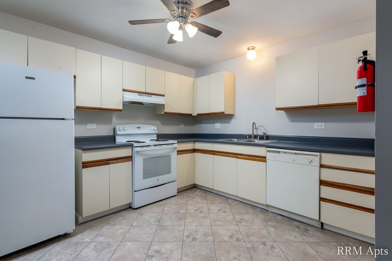 Kitchen with white oak-trimmed cabinets, white appliances, dark counter and a ceiling fan, Pioneer Court apartment 900 1D
