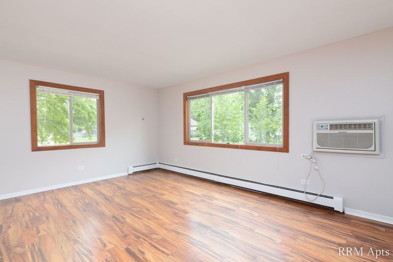 Living room with wood-look floor, two windows with trees and a wall air conditioner, Pioneer Court apartment 900 2A