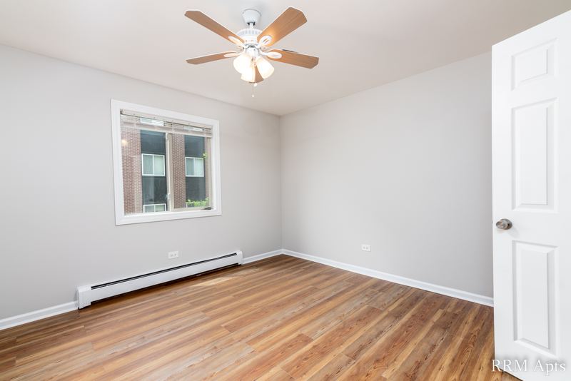 Bedroom with wood-look floor, ceiling fan, window facing a brick building and white door, Pioneer Court apartment 900 2A