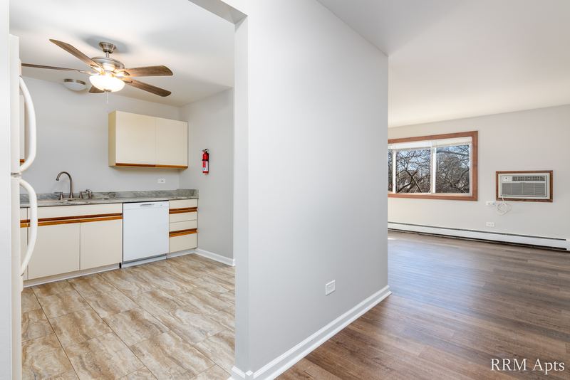 Living room with wood-look floor, window and an open kitchen with white cabinets, Pioneer Court apartment 900-3D