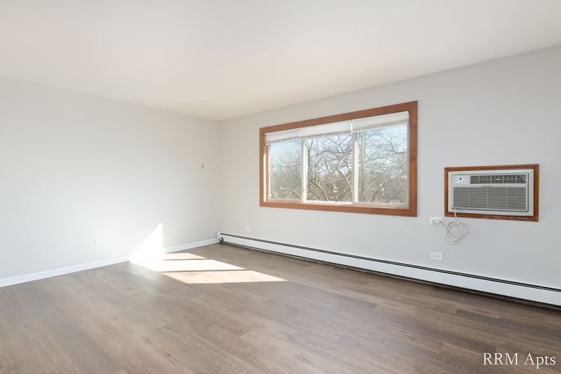 Living room with wood-look floor, window with bare trees and a wall air conditioner, Pioneer Court apartment 900-3D