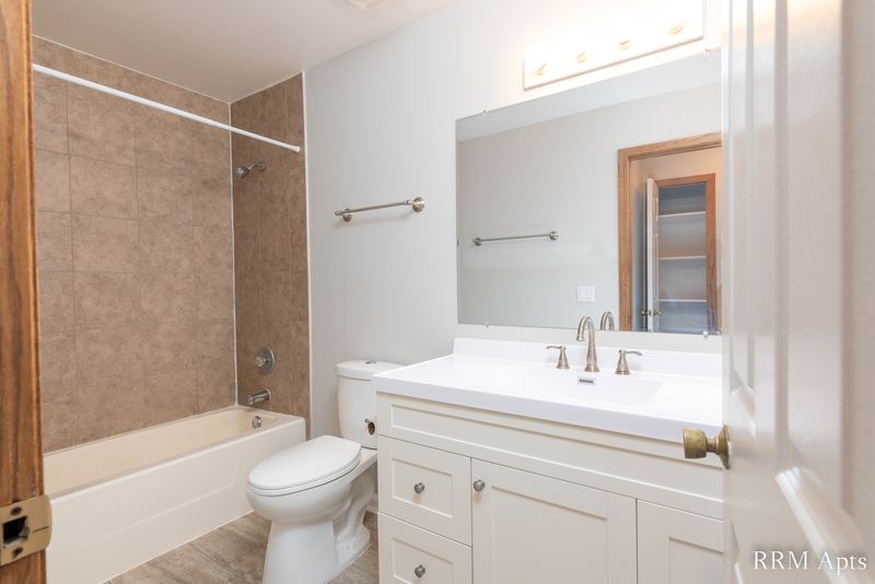 Bathroom with brown tile tub surround, white vanity, mirror, toilet and an oak door, Pioneer Court apartment 900-3D